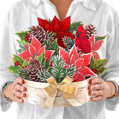 Person holding a decorative Christmas flower arrangement with poinsettias, cardinal, and pinecones.